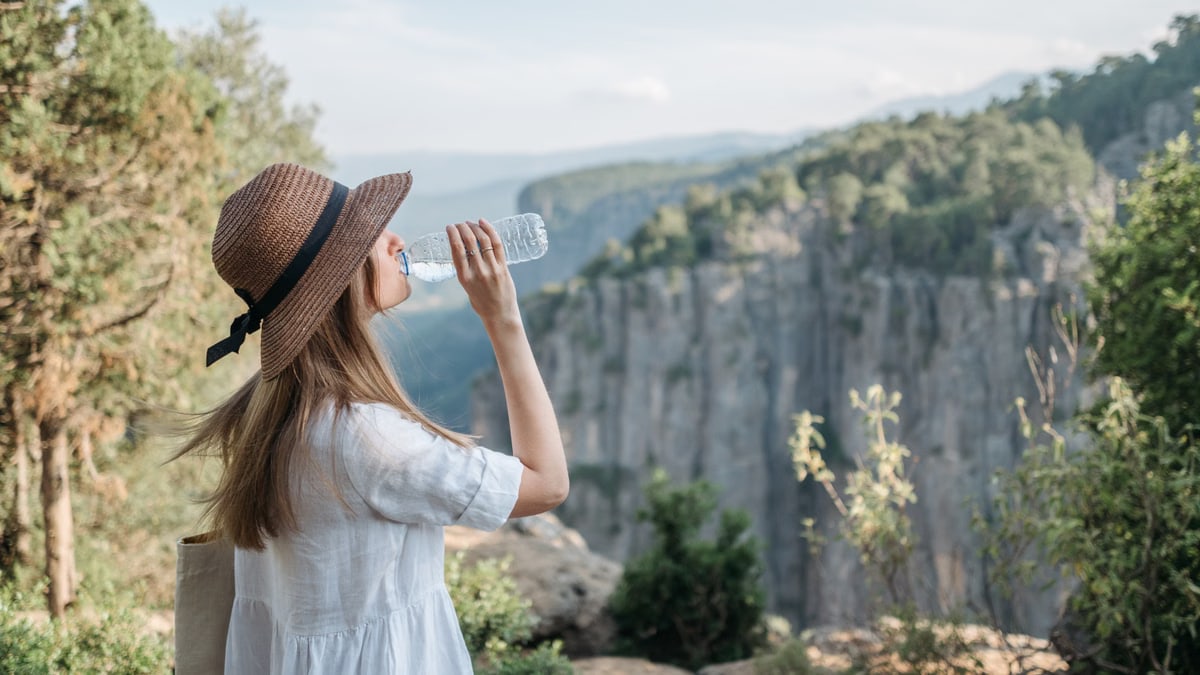 Woman wearing a sun hat drinks water while enjoying a scenic canyon view amidst nature.